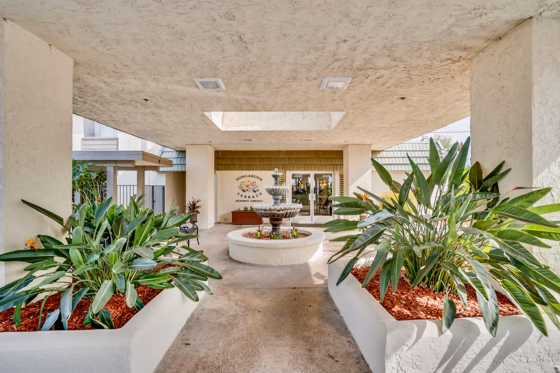 Covered entry to Sungarden Terrace with fountain, planters, and tropical plants.