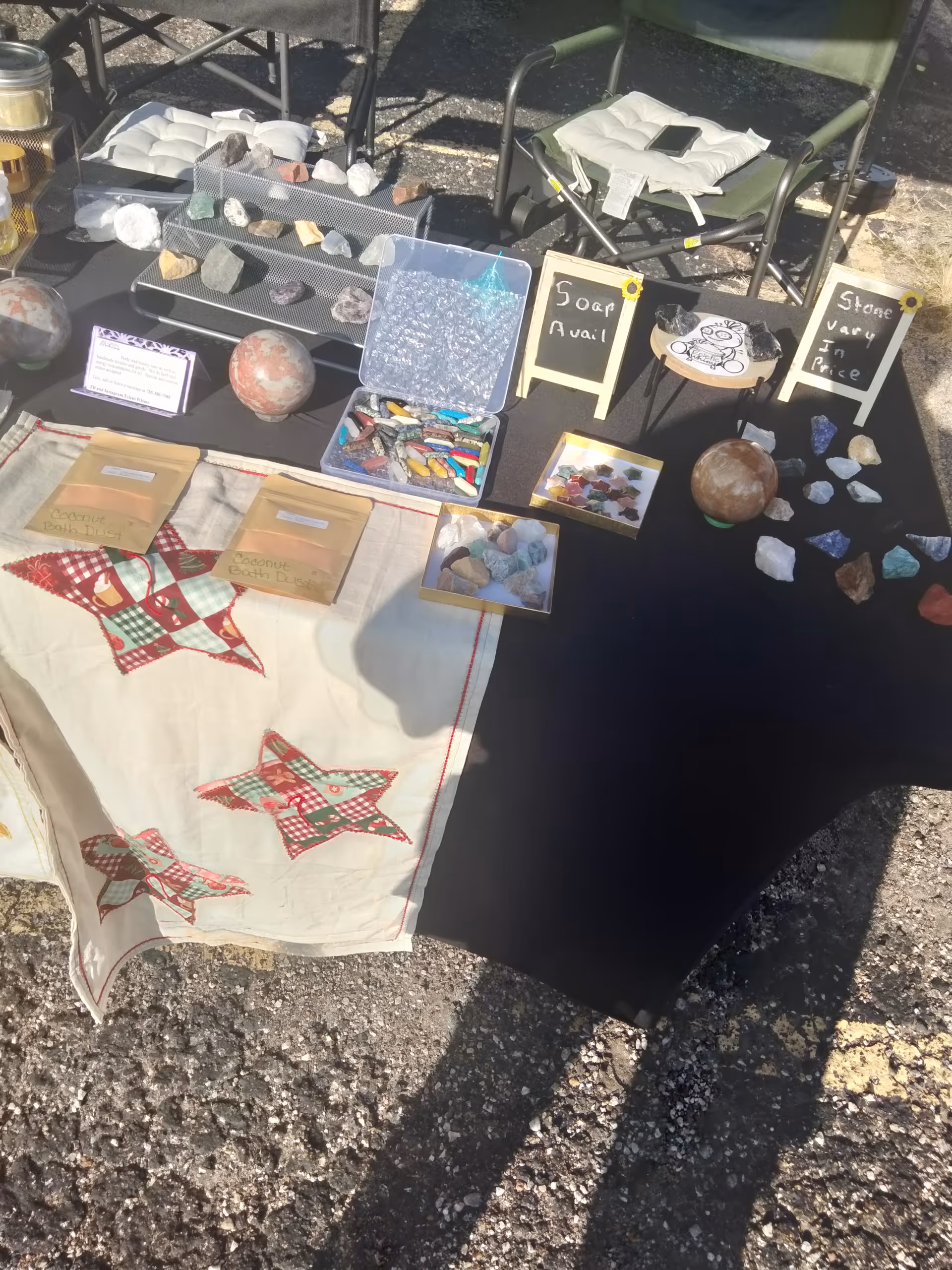 Outdoor market stall displaying rocks, minerals, and crafts on tables with chairs behind.