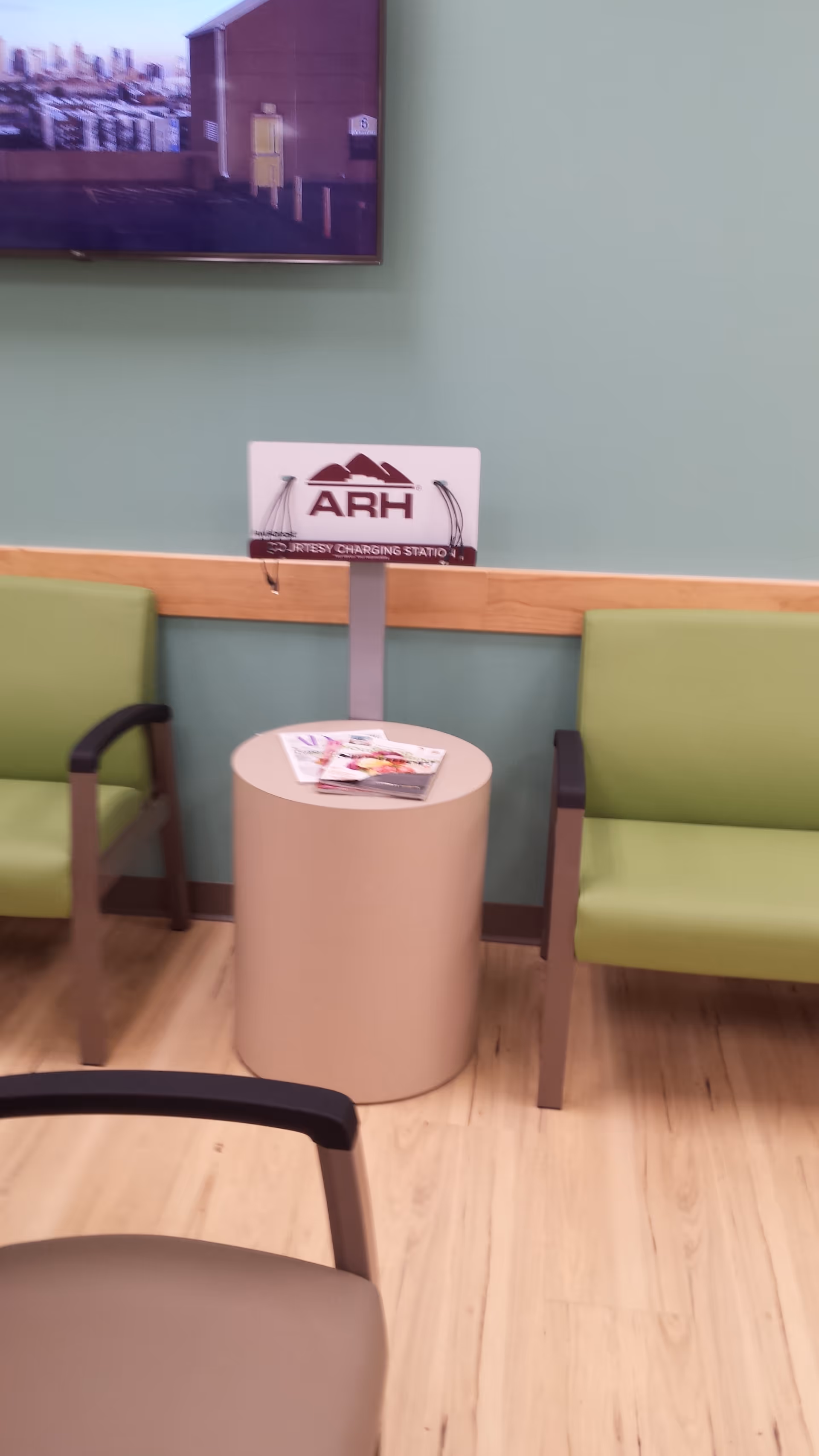 Waiting area with green chairs, a beige round table, magazines, and a wall-mounted screen.