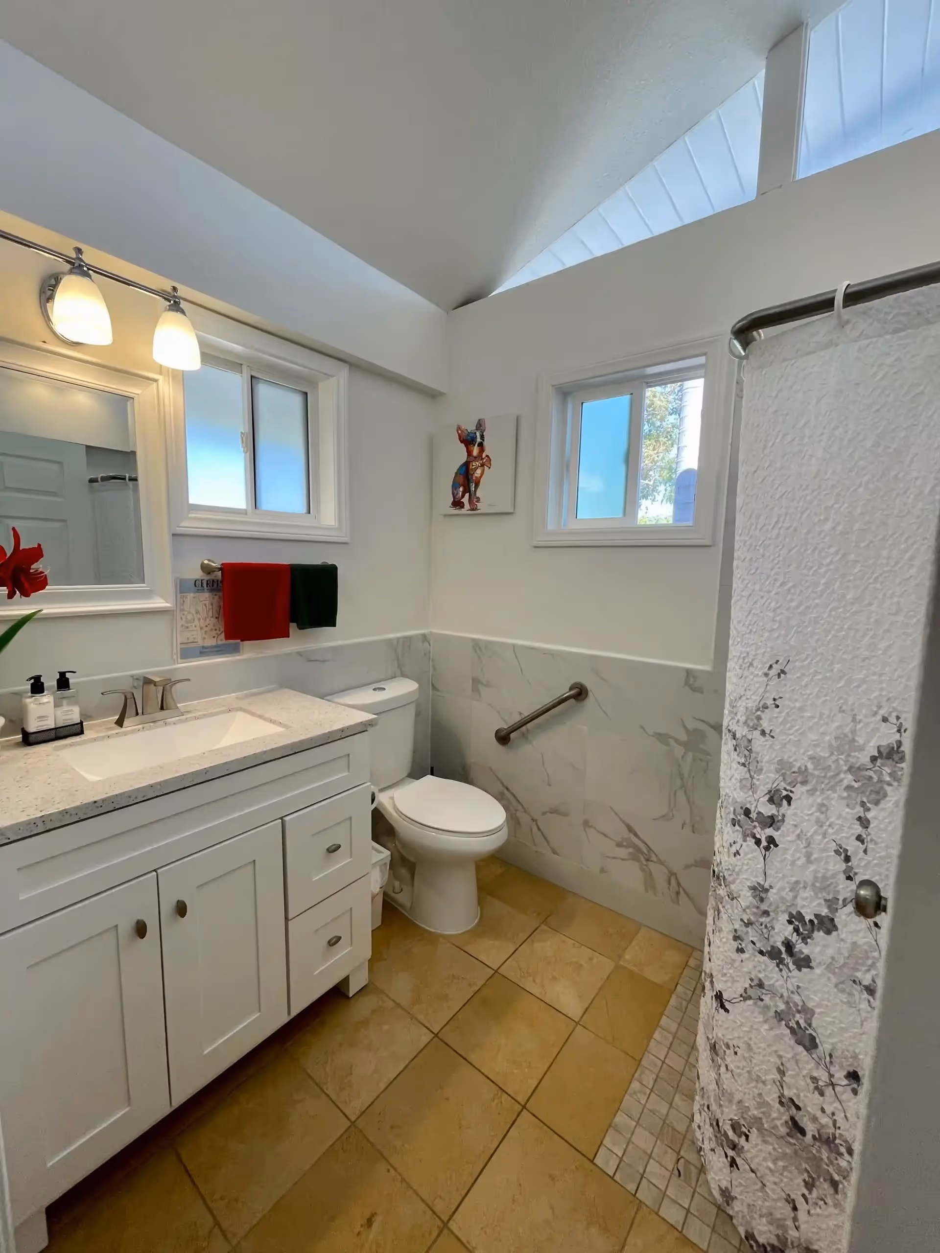 White bathroom with vanity, sink, toilet, shower curtain, grab bar, and two small windows.