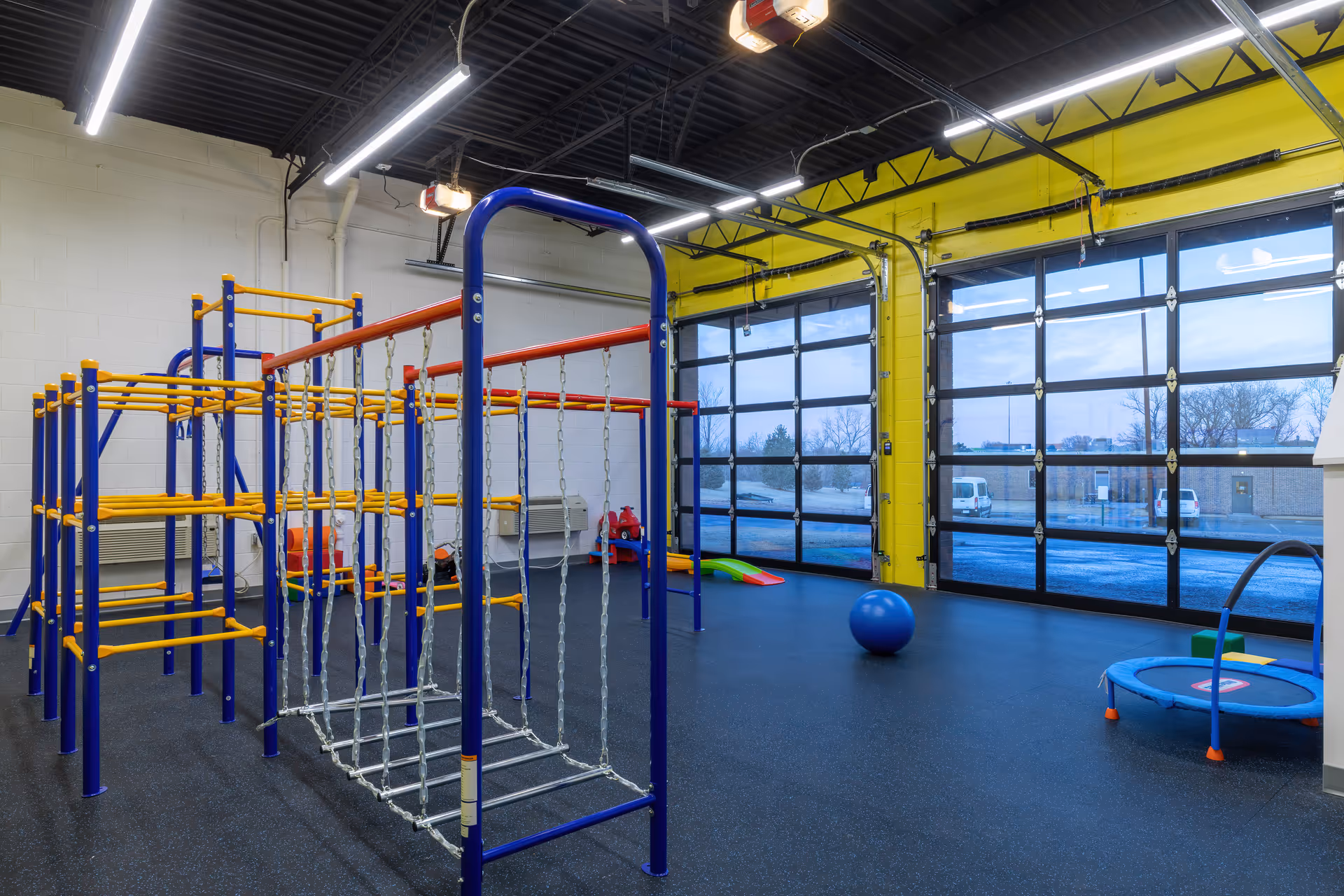 Indoor therapy play area with colorful climbing frames, ropes, and a large glass garage-style door.