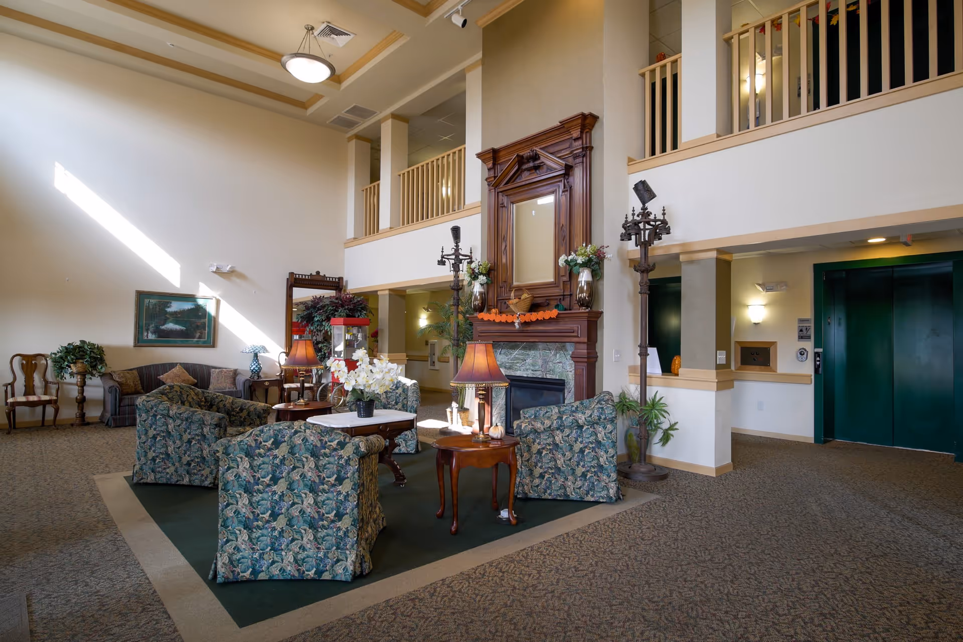 A spacious senior living facility common area with high ceilings and natural light. The room features a seating arrangement with floral patterned armchairs and a sofa around a wooden coffee table. There is a large wooden fireplace with a mirror above it, decorated with flowers and a small pumpkin garland. To the right, there is a green elevator door and a coat rack. The upper level has a balcony with wooden railings overlooking the area.