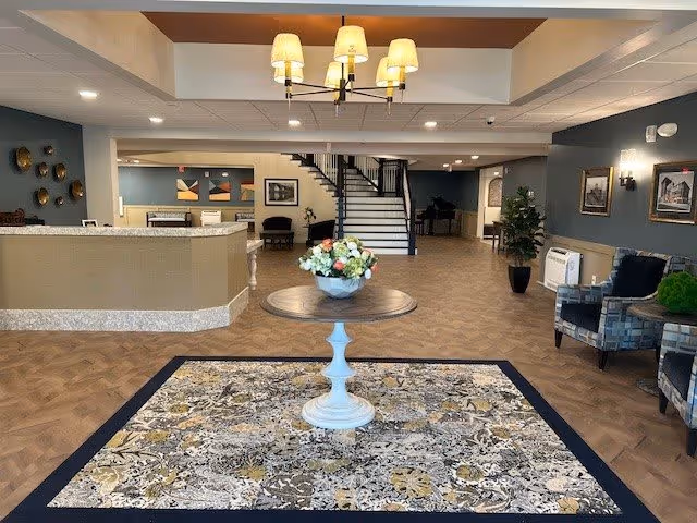 Bright lobby interior with a central round table and flower arrangement on a patterned rug, reception desk to the left, seating to the right, and a staircase in the background.