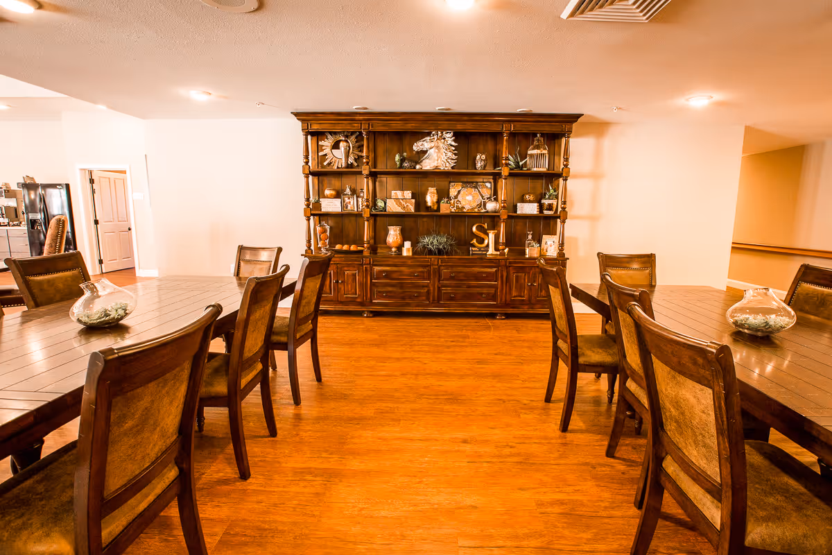 A warm, inviting dining area with two large wooden tables surrounded by chairs. A large wooden cabinet with decorative items such as vases, plants, and sculptures is against the far wall. The room has wooden flooring and soft lighting from ceiling fixtures.