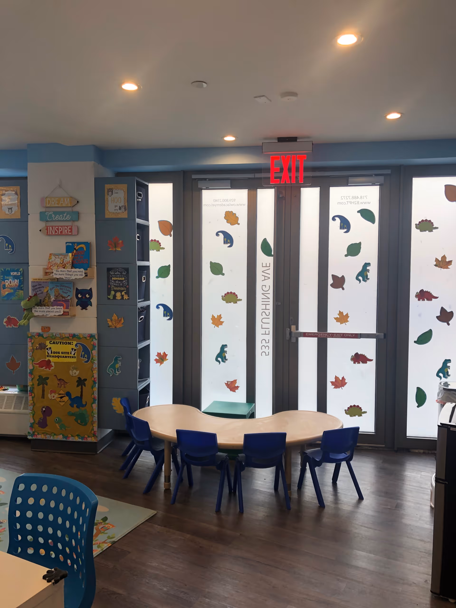 Bright children's playroom with small table, blue chairs, shelves, and leaf-decorated glass doors.