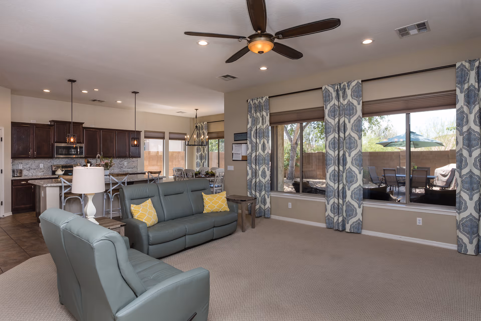 A spacious living room with two green leather sofas, yellow patterned throw pillows, a white table lamp, and large windows with blue and white patterned curtains. The room opens into a kitchen with dark wood cabinets, granite countertops, and a dining area with a table and chairs. Outside the windows, there is a patio area with outdoor furniture and an umbrella.