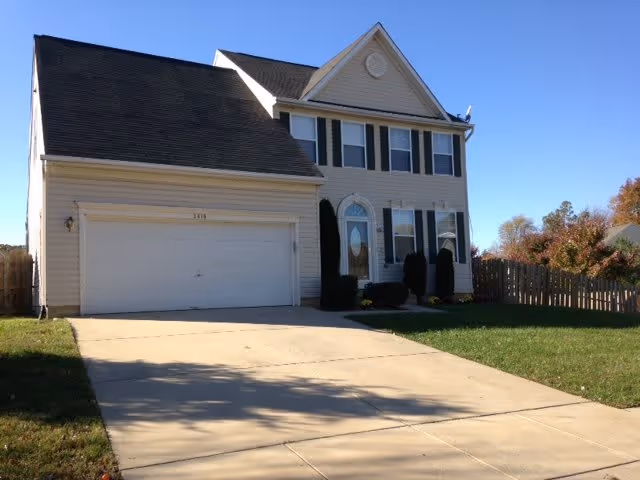 A two-story beige house with black shutters, a double garage, and a concrete driveway. The house has a small front lawn with some bushes and a wooden fence on the right side. The sky is clear and blue.