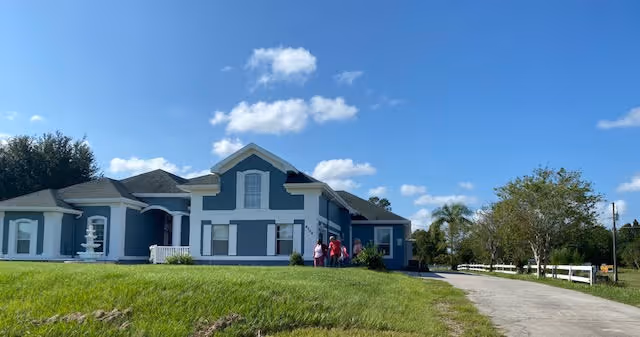 Blue single-story senior housing building with white trim, driveway, and a grassy front yard.