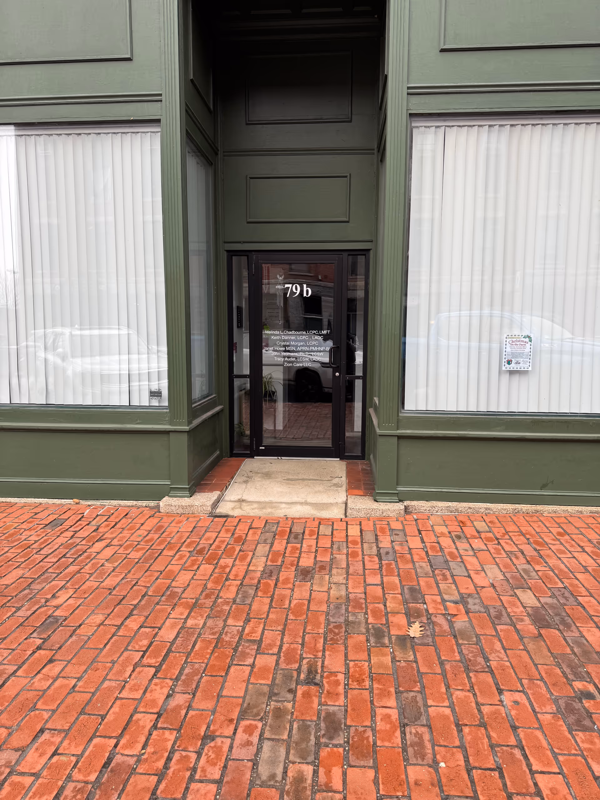 Green brick building exterior with glass door entrance labeled '79b' and brick sidewalk.