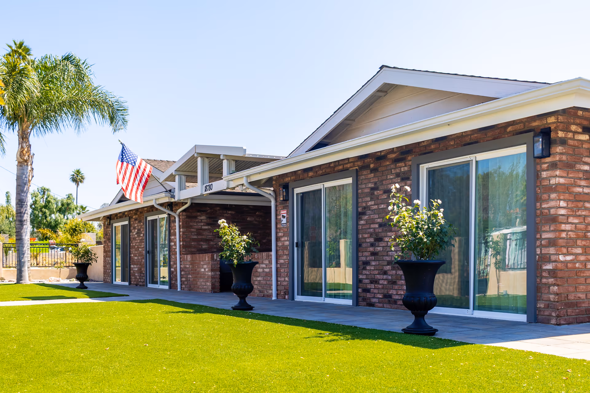 Exterior view of a single-story brick building with large sliding glass doors, potted plants, a well-maintained green lawn, and an American flag mounted near the entrance under a clear blue sky.