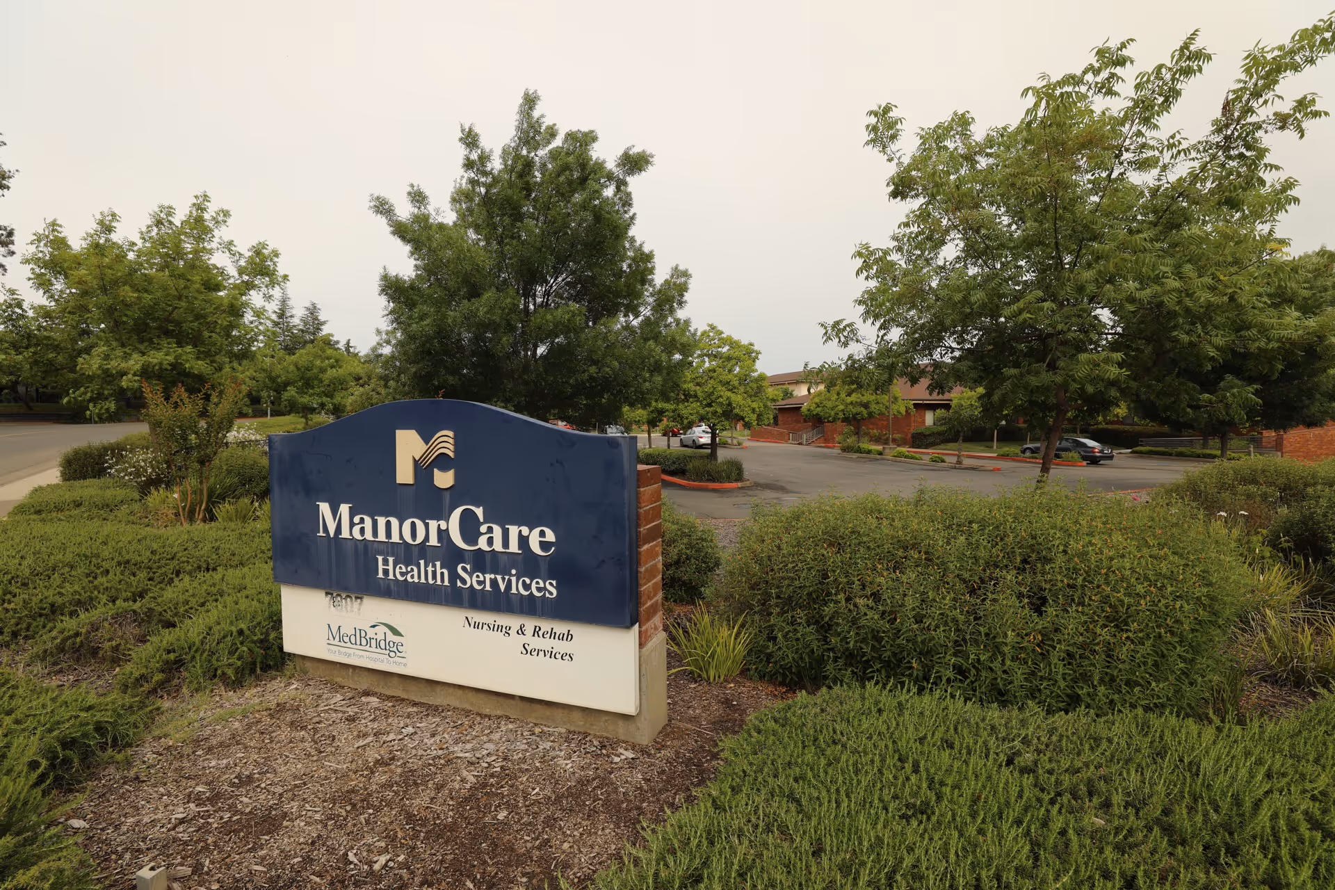 Blue ManorCare Health Services sign sits among landscaping with a parking lot and trees.