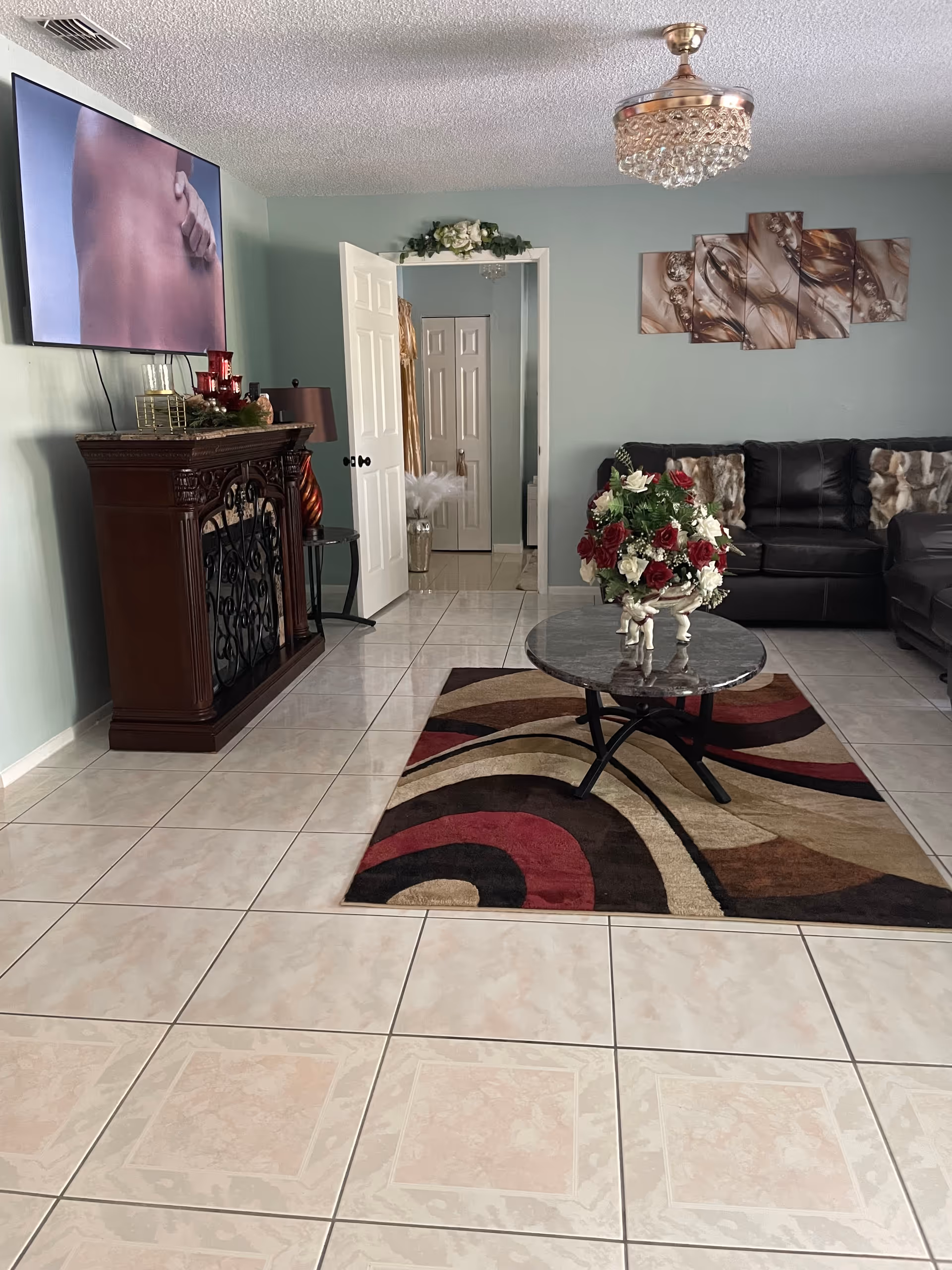 Living room with tiled floor, wall-mounted TV, dark ornate cabinet, and glass coffee table.