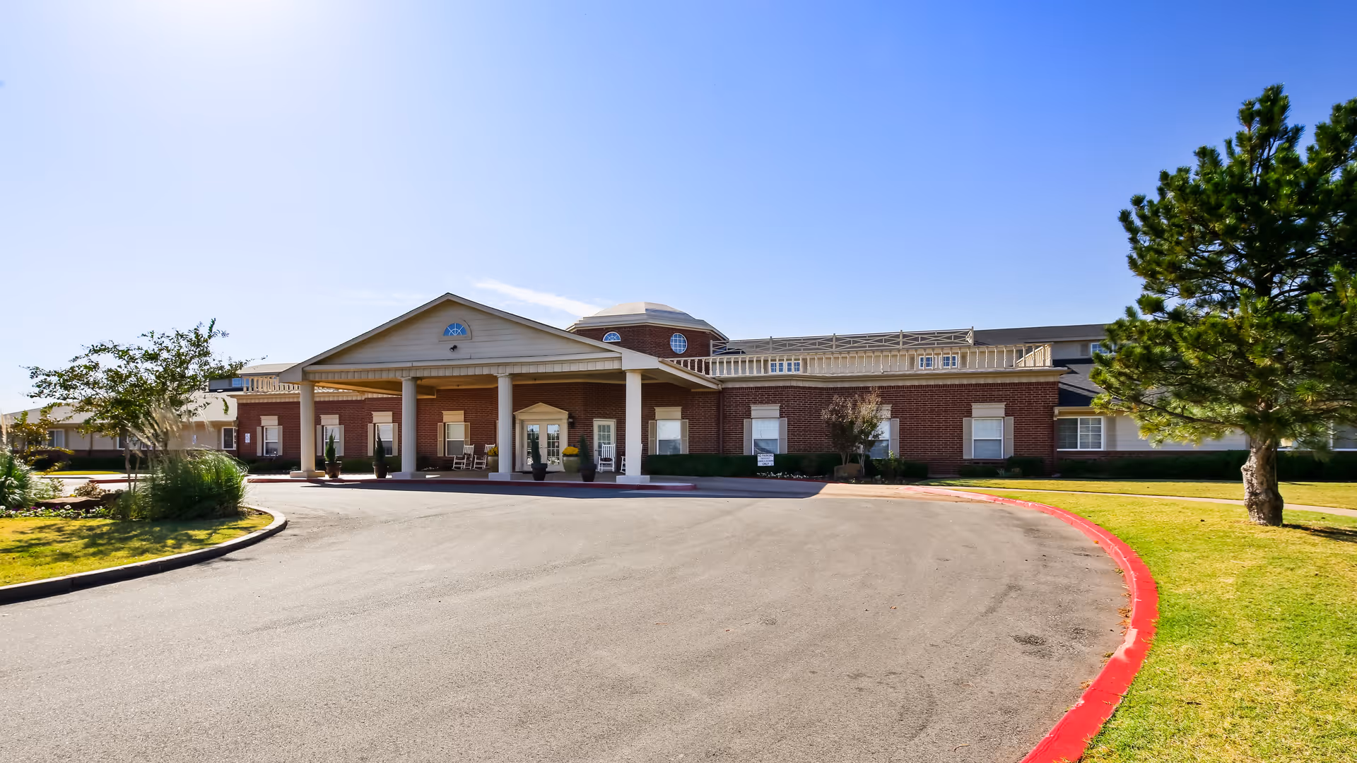 Front exterior view of a single-story brick building with a covered entrance supported by columns, surrounded by a paved driveway and green lawn with trees under a clear blue sky.