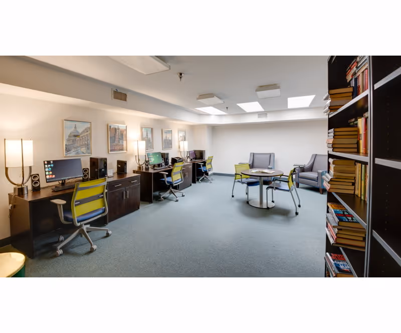 A computer study area with desks, monitors, chairs, and a bookshelf-filled wall.