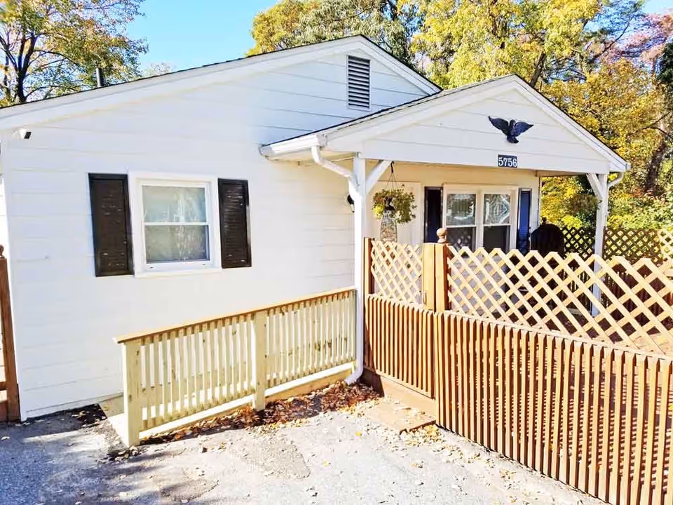 Exterior view of a small white building with black shutters on the windows, a wooden ramp leading to the entrance, and a wooden lattice fence enclosing a porch area. The building has a triangular roof with a black eagle decoration and the number 5756 displayed above the porch. Trees with autumn foliage are visible in the background.