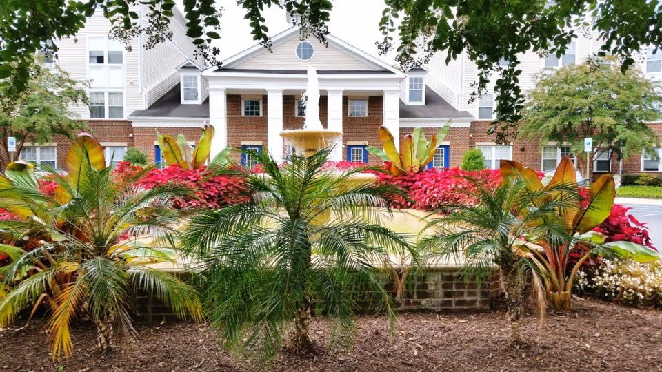 Front entrance of a senior living building with a central fountain, vibrant landscaping, and a brick facade with white columns.