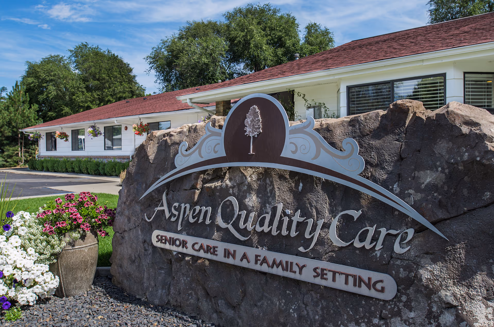 Exterior view of Aspen Quality Care sign on rock with a white building and flowers.