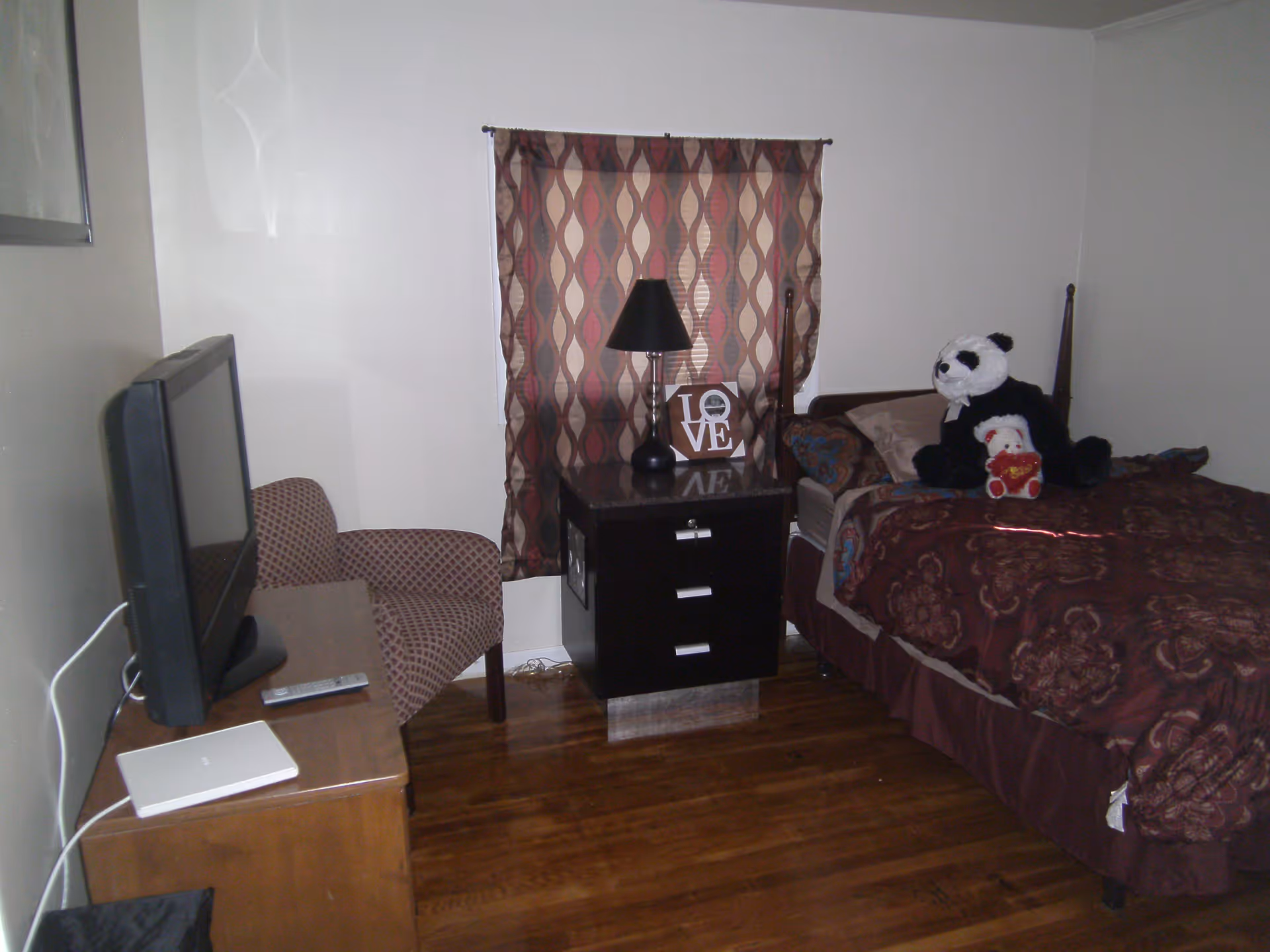 Small bedroom with a bed and stuffed panda, nightstand and lamp, a TV on a dresser, and an armchair.