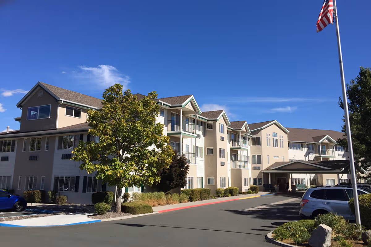 Three-story senior living building with an entrance canopy, parked cars, a tree in front, and an American flag against a clear blue sky.