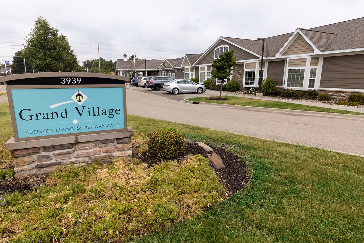 Entrance sign reading 'Grand Village Assisted Living & Memory Care' in front of a single-story assisted living building and parking lot.