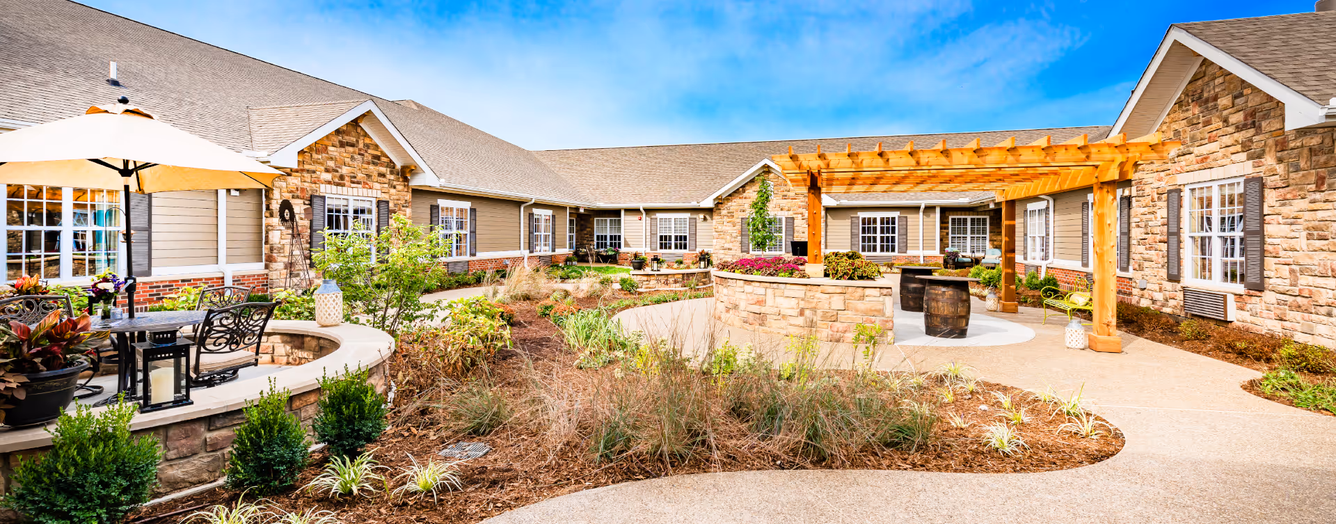 Outdoor courtyard area of a senior living facility with stone and siding exterior walls, a wooden pergola, landscaped garden beds, patio seating with a table and umbrella, and decorative plants under a clear blue sky.