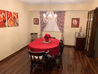 Dining room with a round table covered in a red tablecloth, wooden chairs, chandelier, curtains and hardwood floor.