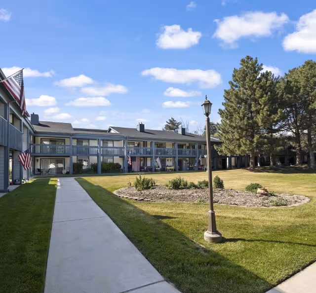 Two-story senior living facility exterior with a walkway, lawn, trees, and American flags.