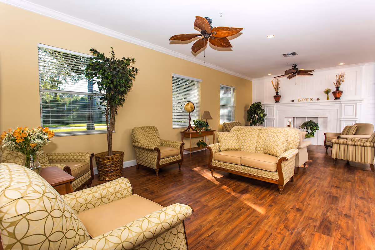 Sunlit communal living room with patterned sofas and armchairs, hardwood floors, potted plants, ceiling fans, and a white mantel fireplace.