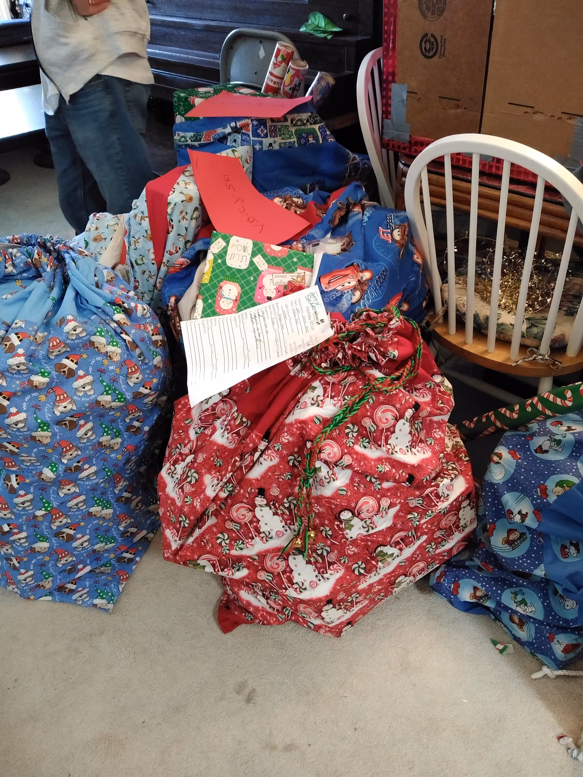 Colorful wrapped gift bags and boxes piled on the floor beside chairs in a cluttered room.