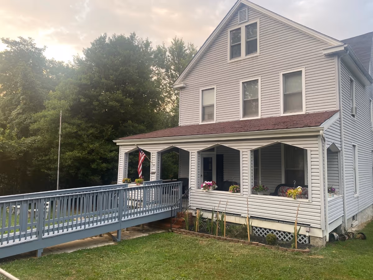 Two-story gray building with a wraparound porch, ramp, and flower planters.