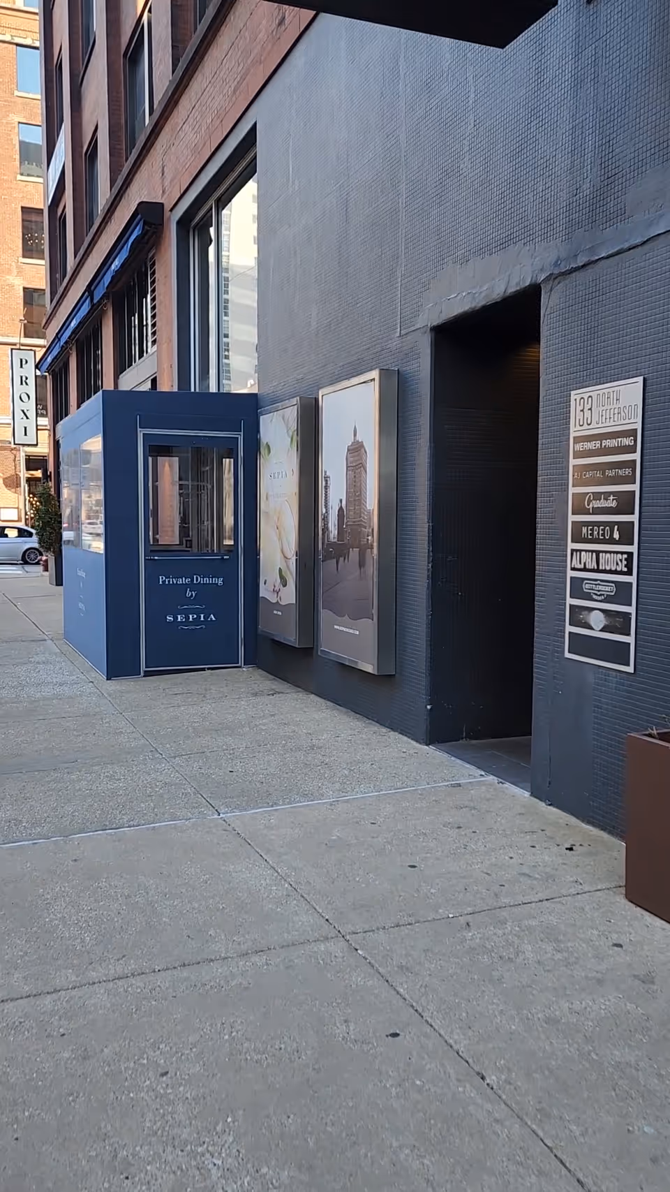 Urban sidewalk scene with a dark building exterior, posters, and a blue storefront entrance.