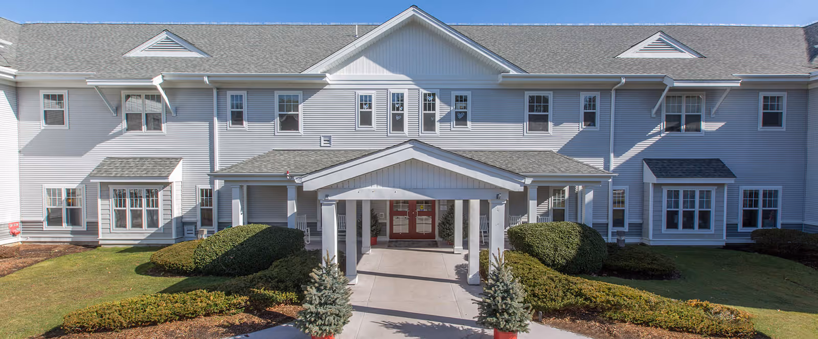Front exterior view of a two-story senior living facility building with a covered entrance, multiple windows, and well-maintained landscaping including bushes and small trees.