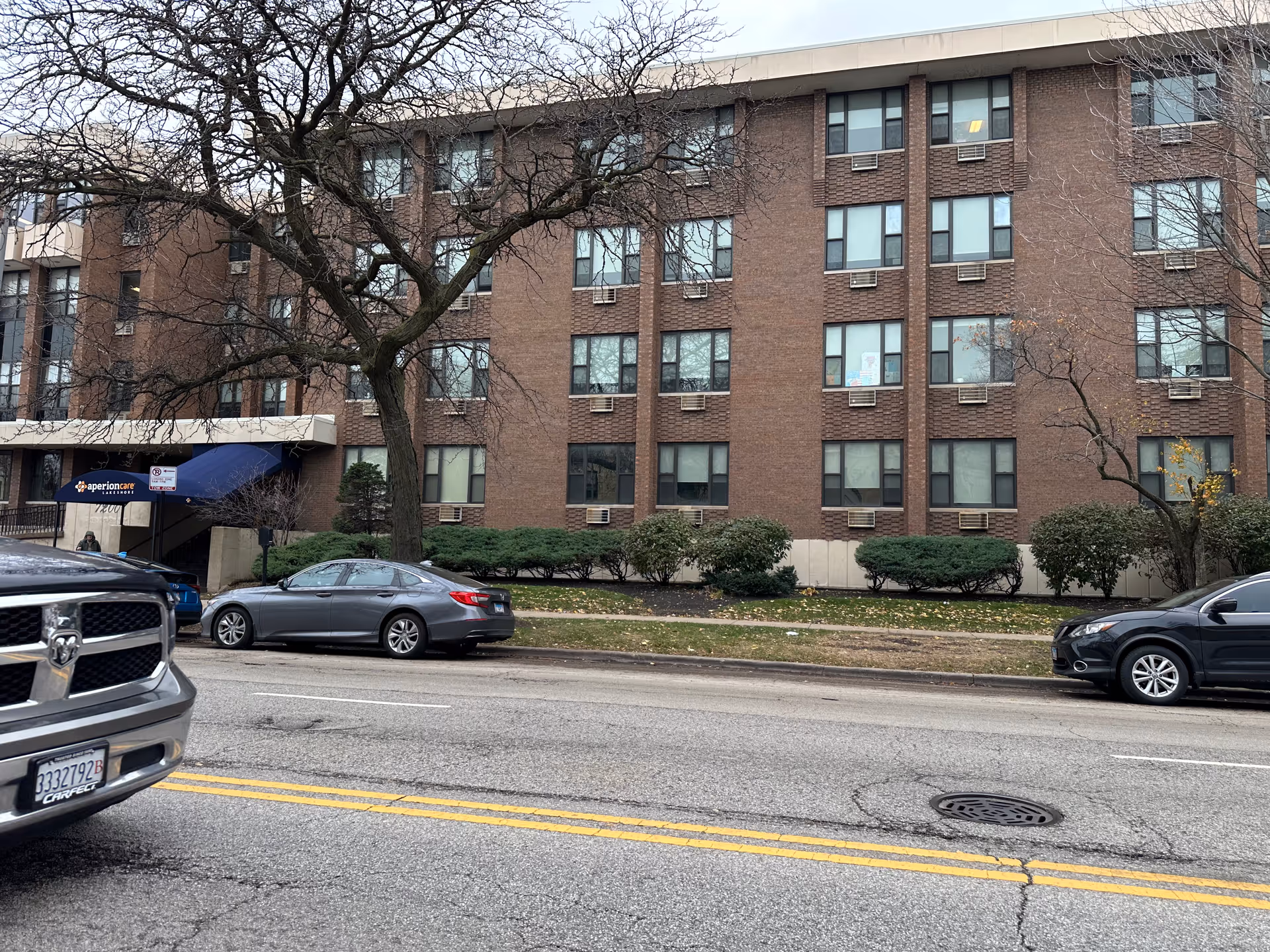 Exterior view of a multi-story brick building with several windows, some with air conditioning units. There are trees without leaves and bushes in front of the building. Several cars are parked along the street in front of the building. A blue awning near the entrance displays the name 'aperioncare Lakeshore'.