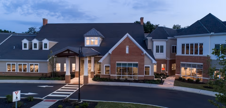 Front exterior of a brick-and-siding senior living facility with a covered entrance and illuminated windows at dusk.