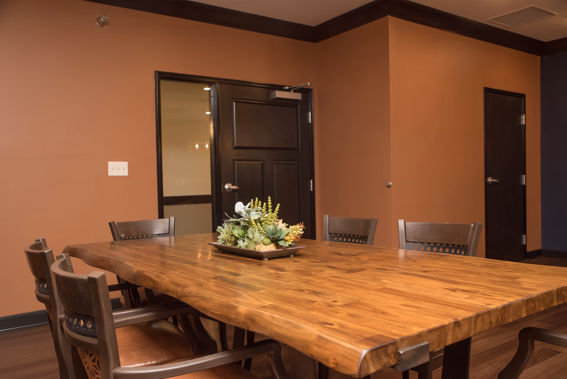A wooden dining table with six chairs around it in a room with brown walls and dark trim. A decorative centerpiece with green and yellow plants is placed on the table. There are two dark-colored doors visible in the background.