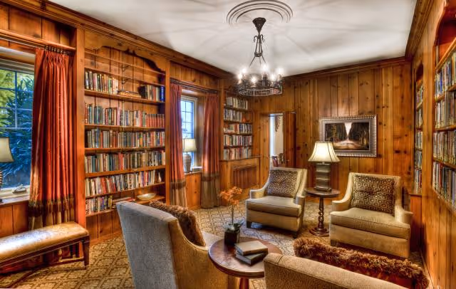 Cozy wood-paneled library-style sitting room with bookshelves, armchairs, side tables, and a chandelier.