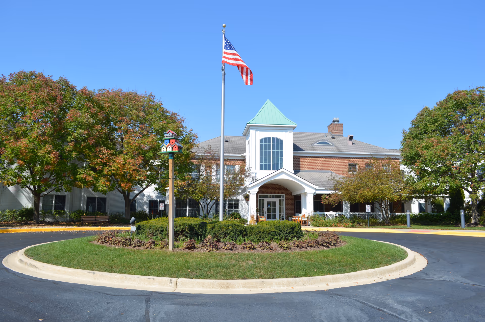 Front view of a senior living facility with circular driveway, flagpole, and trees.