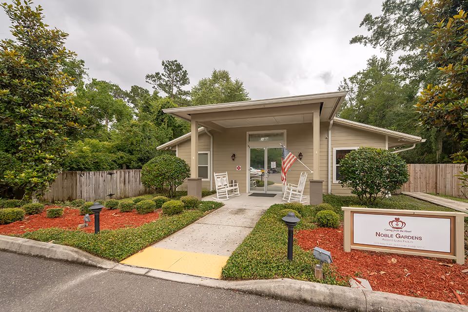 Front entrance of a single-story assisted living building with rocking chairs on the covered porch, an American flag, landscaped shrubs, and a Noble Gardens sign.