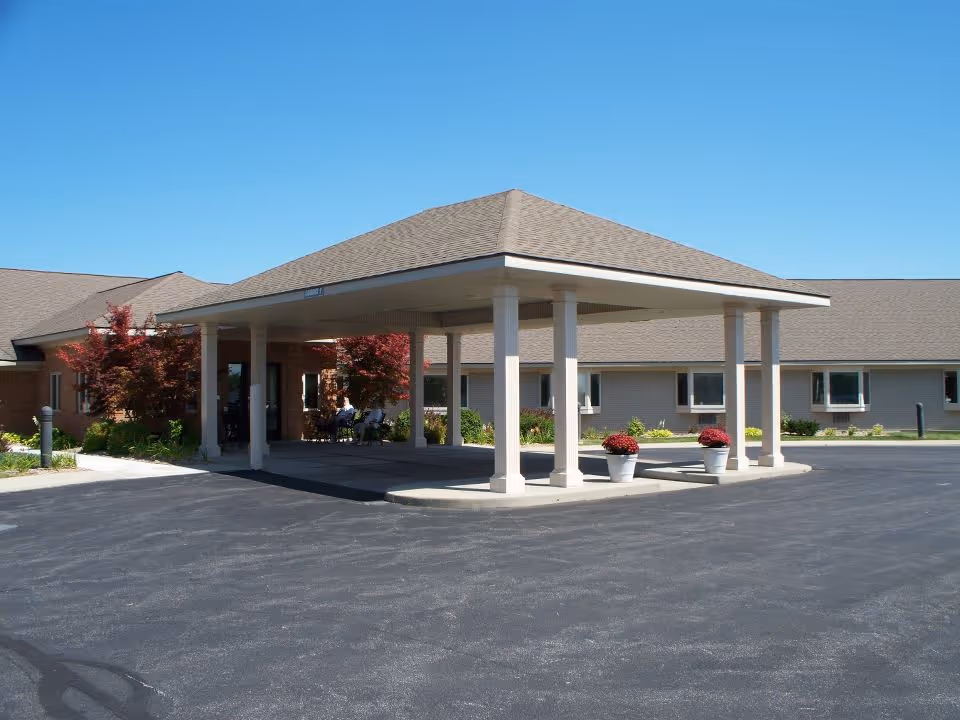 Exterior view of the entrance to Edgewood Assisted Living Center featuring a covered drop-off area with white pillars, two potted plants with red flowers, and a paved driveway under a clear blue sky.
