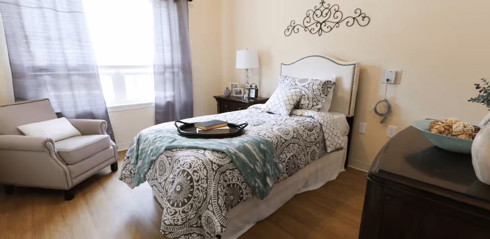 A cozy bedroom with a single bed covered in patterned bedding and a teal throw blanket. There is a beige armchair with a white pillow near a window with gray curtains. A dark wooden nightstand with a lamp and picture frames is beside the bed. A decorative metal wall art piece hangs above the headboard. A dark wooden dresser with a bowl of decorative items is visible in the foreground.