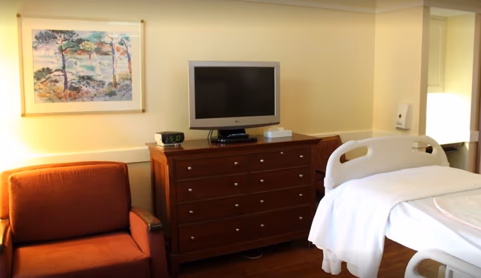 Hospital-style patient room with a bed, dresser, TV, and orange chair.
