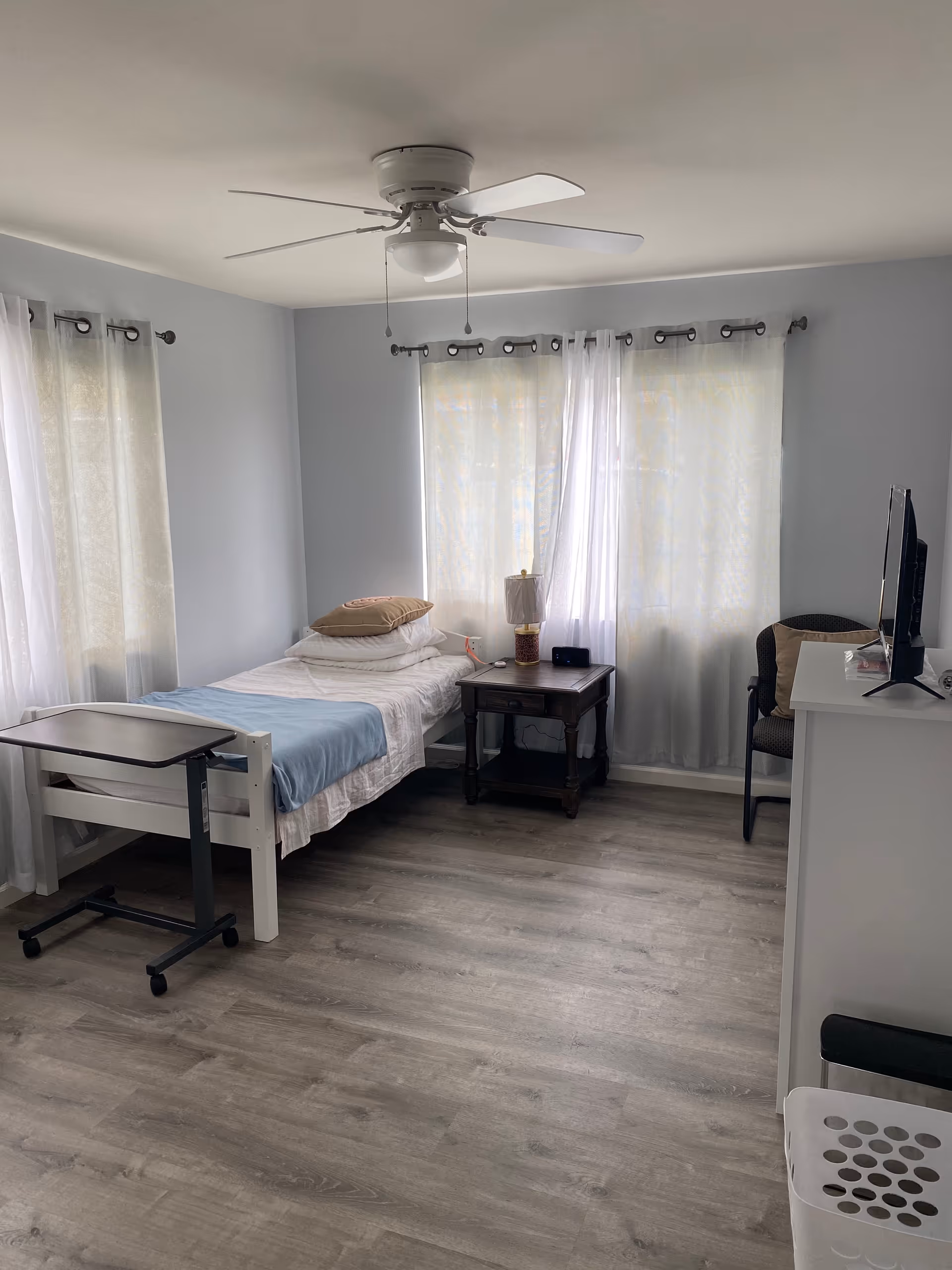 Simple furnished bedroom with single bed, nightstand, dresser, TV, ceiling fan, curtains.