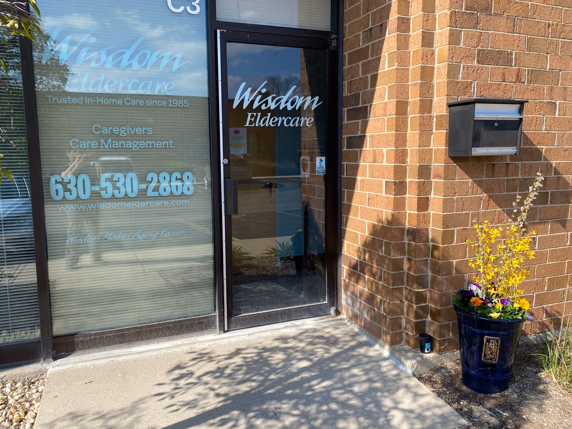 Glass entrance door on a brick Wisdom Eldercare building with a flower planter.
