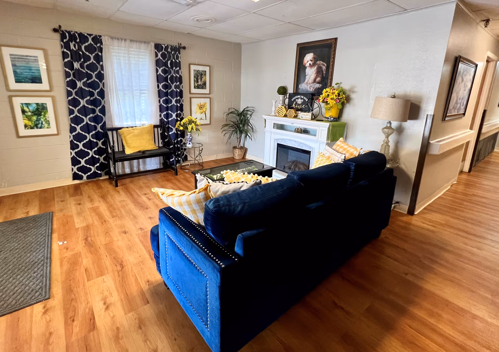 A cozy living room area with a blue couch adorned with yellow and white pillows, a white fireplace decorated with flowers and a framed picture of a dog above it. There is a black bench with a yellow pillow in front of a window with navy blue and white patterned curtains. The room has wooden flooring and light-colored walls with several framed pictures. A lamp stands near the couch and a hallway is visible to the right.