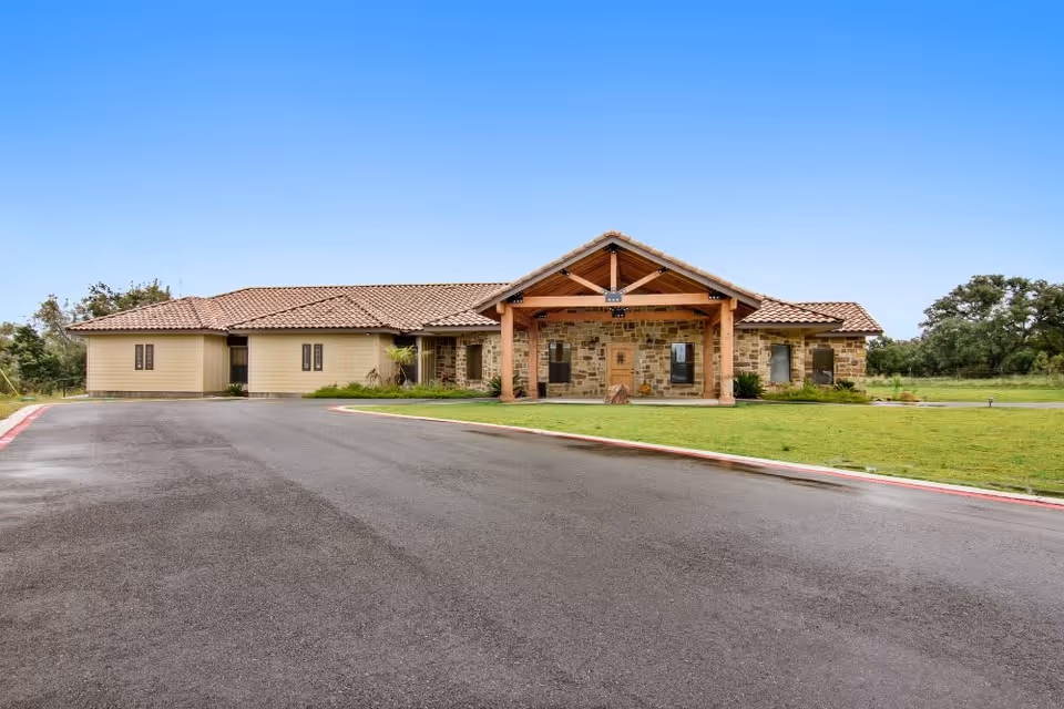 Front exterior view of a single-story building with a stone and beige siding facade, a tiled roof, and a covered entrance supported by wooden beams. The building is surrounded by a paved driveway and green lawn under a clear blue sky.