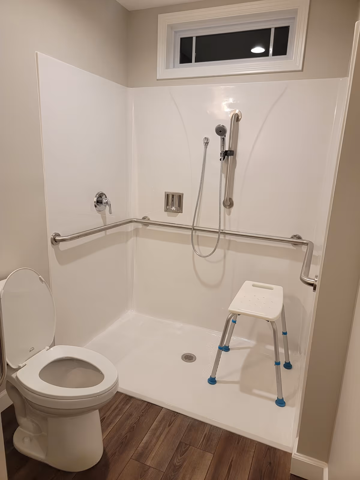 Accessible walk-in shower with grab bars, a small white stool, and a toilet in beige bathroom.