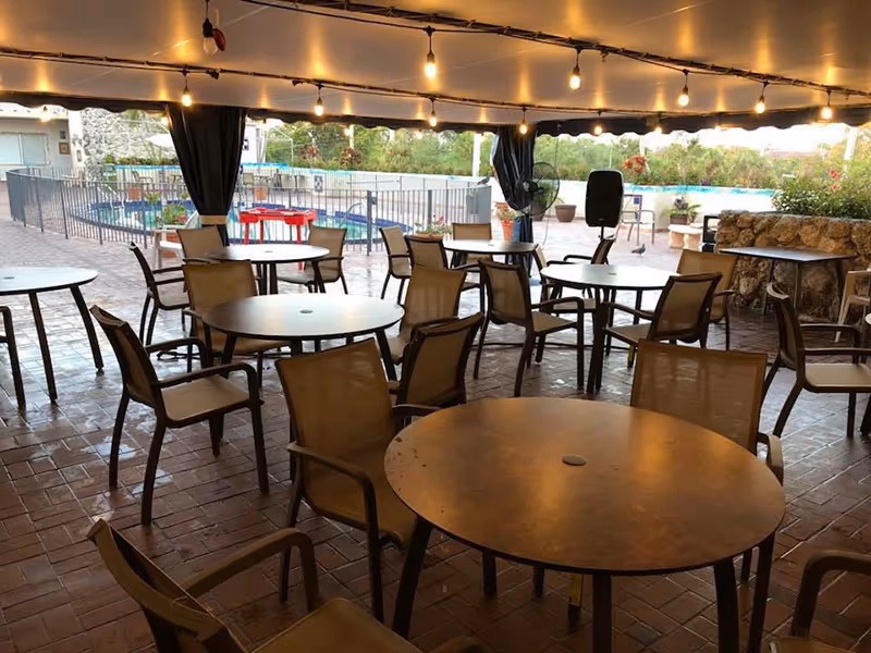 Covered outdoor seating area with multiple round tables and chairs arranged on a brick floor. String lights hang from the ceiling, and beyond the seating area, there is a fenced swimming pool and greenery in the background.