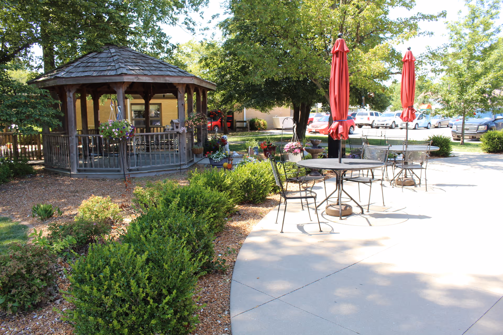 Outdoor patio area with metal tables and chairs, each table having a closed red umbrella. There is a wooden gazebo surrounded by greenery and flowers, with trees providing shade. In the background, there is a parking lot with several cars.