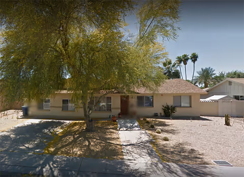 Single-story beige house with driveway, tree in front yard, and desert landscaping.