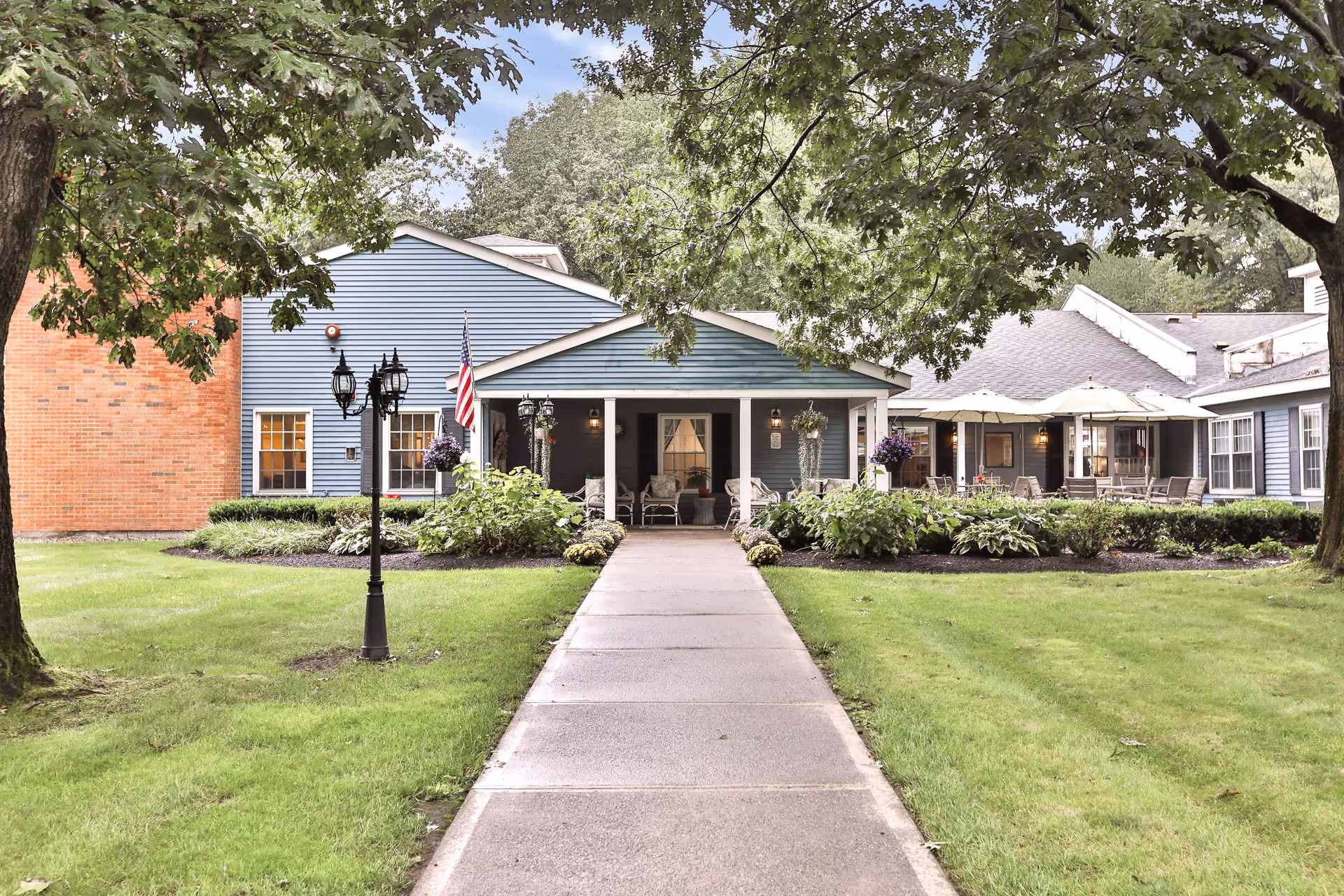 Front exterior view of Peregrine Senior Living at Crossgate showing a pathway leading to the entrance with a covered porch, surrounded by green lawns, trees, shrubs, and outdoor seating with umbrellas.